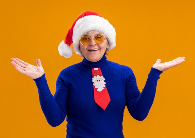 Free photo smiling elderly woman in sun glasses with santa hat and santa tie holding hands open isolated on orange background with copy space