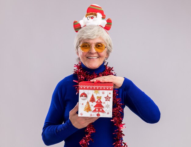 サンタのヘッドバンドと首の周りの花輪とサングラスで笑顔の年配の女性は、コピースペースで白い壁に分離されたクリスマスギフトボックスを保持します。