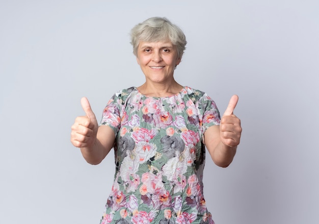 Улыбающаяся пожилая женщина показывает палец вверх двумя руками, изолированными на белой стене