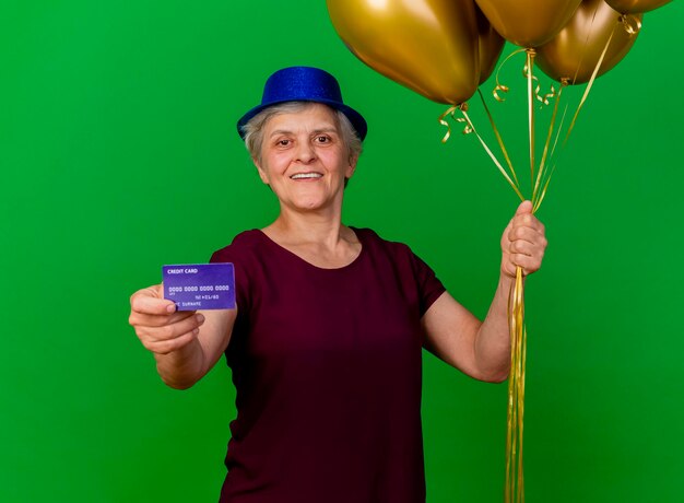 Smiling elderly woman wearing party hat holds credit card and helium balloons on green