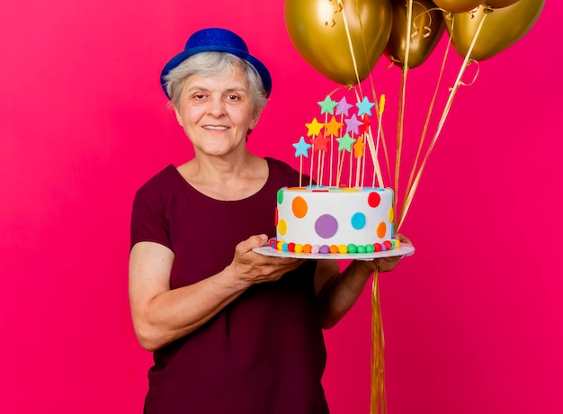 Smiling elderly woman wearing party hat holds helium balloons and birthday cake looking at camera on pink