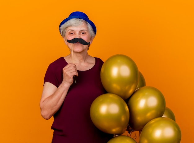 La donna anziana sorridente che porta il cappello del partito tiene i palloni dell'elio e i baffi falsi sul bastone isolato sulla parete arancione