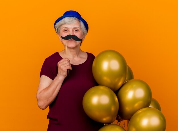 Smiling elderly woman wearing party hat holds helium balloons and fake mustache on stick isolated on orange wall