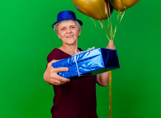 Smiling elderly woman wearing party hat holds helium balloons and gift box on green
