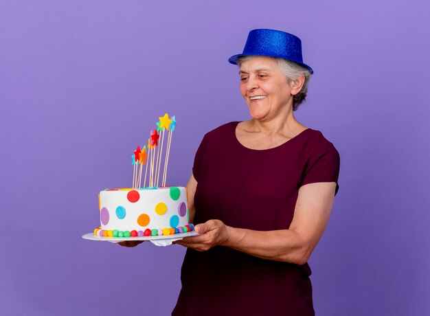 Smiling elderly woman wearing party hat holds and looks at birthday cake isolated on purple wall with copy space