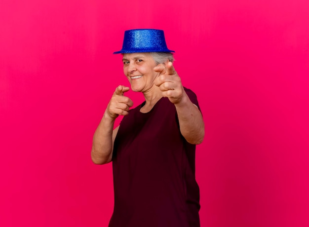 Foto gratuita sorridente donna anziana indossando il cappello da festa guarda e punta alla telecamera con due mani sul rosa