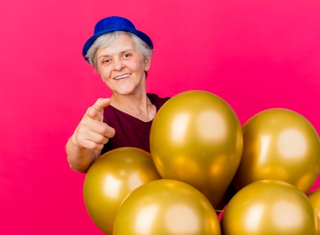 Smiling elderly woman wearing party hat stands behind of helium balloons pointing at camera on pink