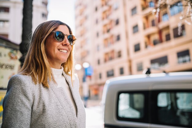 車の近くの通りにサングラスをかけたエレガントな若い女性の笑みを浮かべてください。