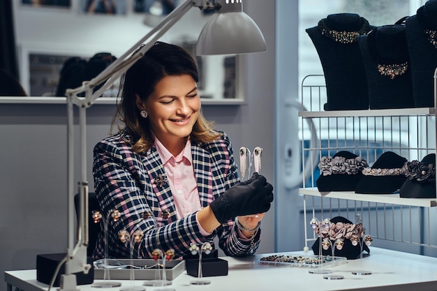 Smiling elegantly dressed female jewelry worker showing beautiful precious earrings with gemstones in a luxury jewelry store.