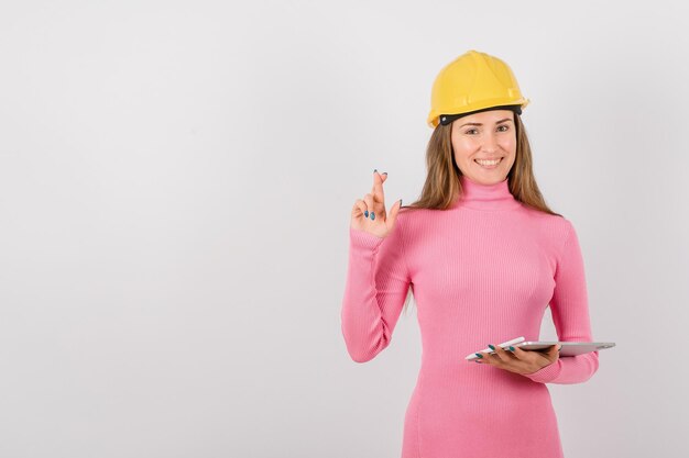 Smiling engineer girl is crossing her fingers by holding tablet computer on white background