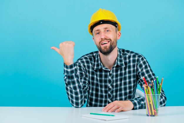 Smiling engineer man is pointing left with thumb on blue background