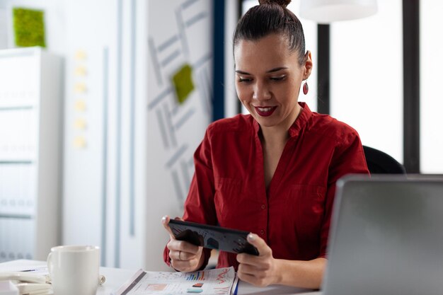 Smiling entrepreneur having a break playing mobile games at the startup office. Employee in red shirt sitting at desk holding smartphone. Businesswoman with gadget streaming content on social media.