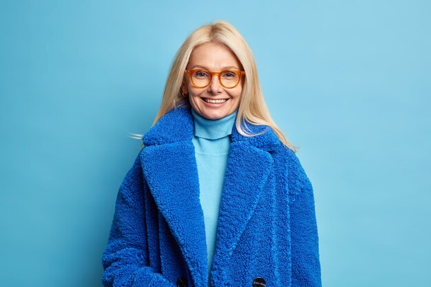 Smiling European woman with blonde hair dressed in blue winter coat has happy mood wears eyewear.