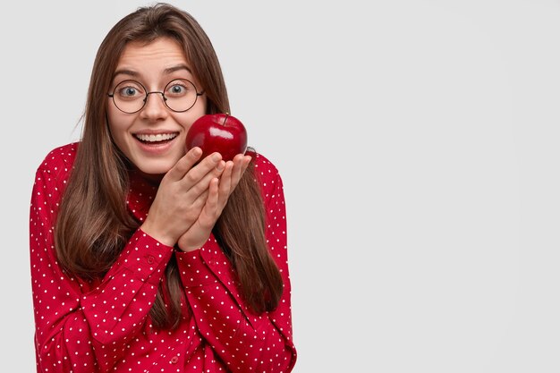 기뻐하는 표정으로 웃는 유럽 여성, 세련된 옷을 입은 빨간 사과 운반, 둥근 안경, 과일 먹는 것을 즐깁니다.