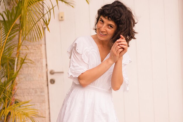 Smiling european young brunette girl wearing white sundress tilts her head looking at camera standing on street near house