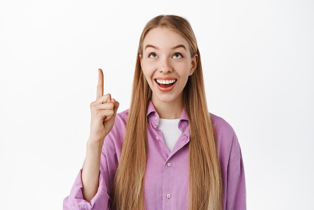 Smiling excited blond girl looking and pointing up with happy face showing advertisement above checking out interesting concept standing over white background