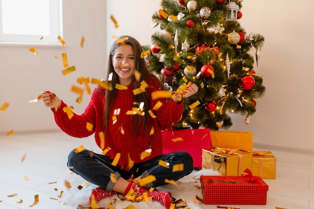 Smiling excited pretty woman in red sweater sitting at home at Christmas tree throwing golden confetti surrounded with presents and gift boxes