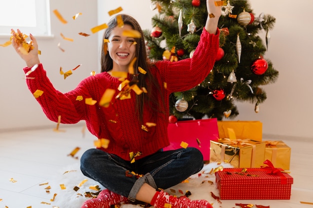 Улыбающаяся возбужденная красивая женщина в красном свитере сидит дома у елки, бросая золотые конфетти в окружении подарков и подарочных коробок