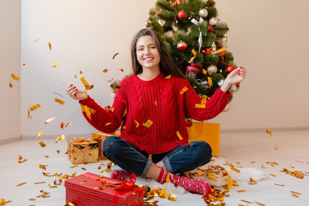 Smiling excited pretty woman in red sweater sitting at home at Christmas tree throwing golden confetti surrounded with presents and gift boxes