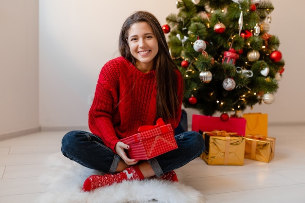 Smiling excited pretty woman in red sweater sitting at home at Christmas tree unpacking presents and gift boxes