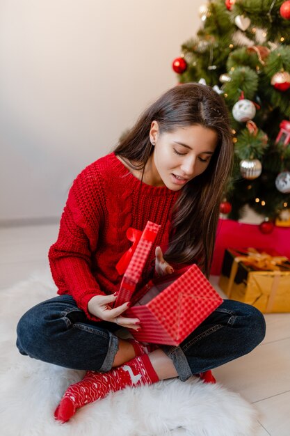 プレゼントやギフトボックスを開梱クリスマスツリーで家に座っている赤いセーターで興奮したきれいな女性の笑顔