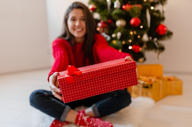 Улыбающаяся возбужденная красивая женщина в красном свитере сидит дома у елки, распаковывая подарки и подарочные коробки
