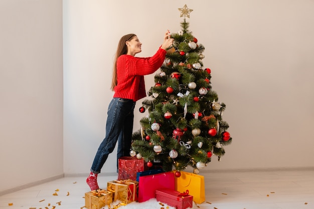 プレゼントやギフトボックスに囲まれたクリスマスツリーを飾る家に立っている赤いセーターで興奮したきれいな女性の笑顔