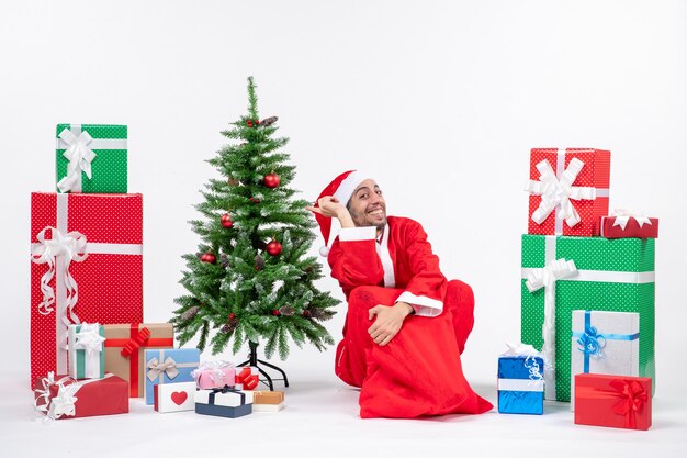 Smiling excited young man dressed as Santa claus with gifts and decorated Christmas tree sitting on the ground on white background