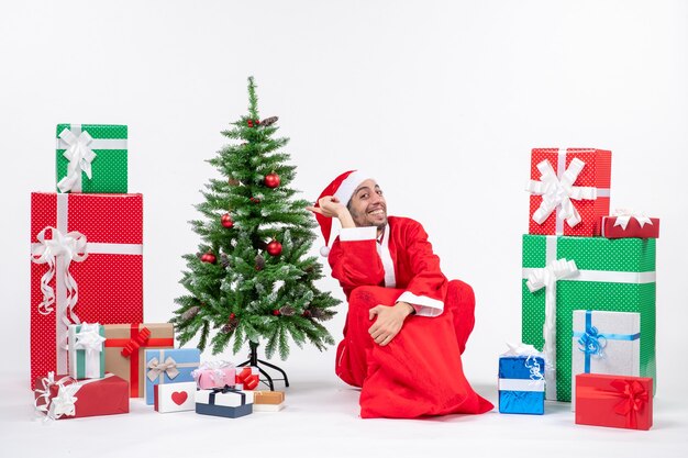 Улыбающийся возбужденный молодой человек в костюме Санта-Клауса с подарками и украшенной елкой сидит на земле на белом фоне