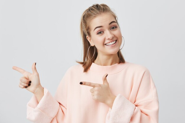 あなたの広告やテキストのコピースペースを指しているピンクのスウェットシャツで金髪の若い女性を笑顔します。何かを宣伝する彼女の白い歯を示す笑顔で肯定的な女の子