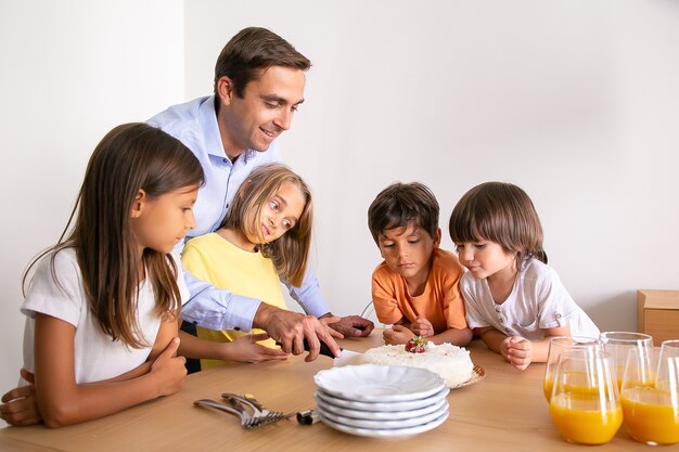 Smiling father cutting tasty birthday cake for children. Lovely little kids standing near table, celebrating birthday together and waiting for dessert. Childhood, celebration and holiday concept