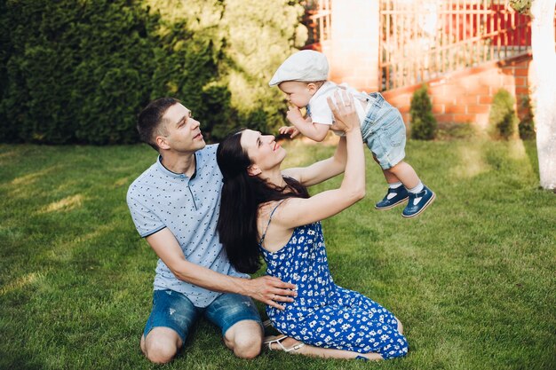 Smiling father and mother playing with their son while spending weekend together and sitting on the lawn in the backyard of the house