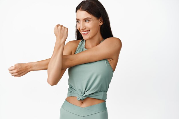 フィットネスジムのエクササイズの白い背景のためのトレーニングウォームアップの前に腕を伸ばしている女性アスリートの笑顔