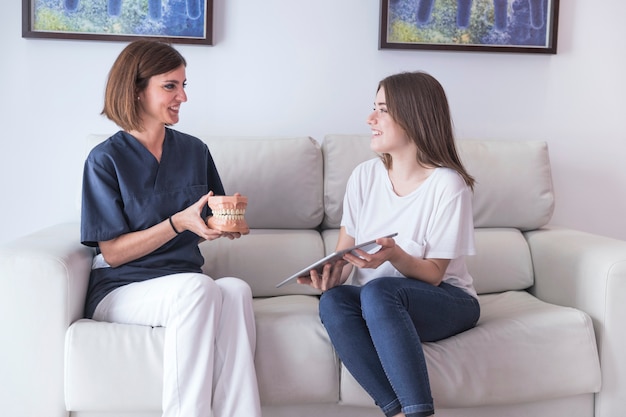 Smiling female dentist showing teeth model to patient holding digital tablet