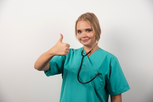 Smiling female doctor making thumbs up on white wall. 