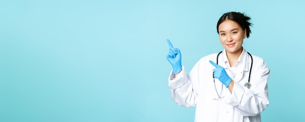 Smiling female doctor physican in medical uniform and sterile gloves pointing fingers left at promo