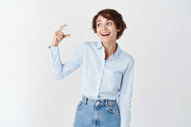 Smiling female entrepreneur showing small size, looking at fingers making little gesture, standing on white wall