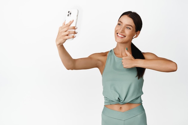 Smiling female fitness instructor record sport video showing thumbs up while taking selfie on mobile phone wearing activewear white background