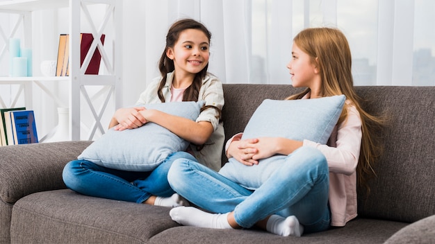 Smiling female friends sitting on sofa with cushion looking to each other