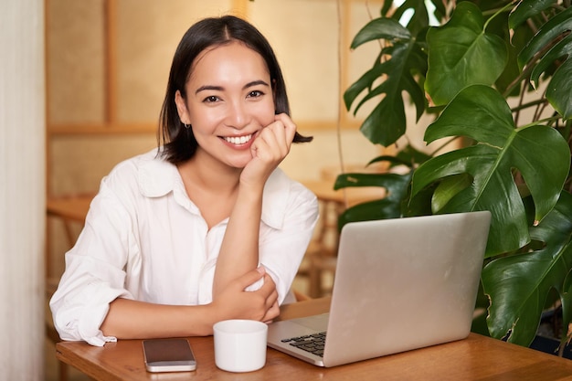 無料写真 笑顔の女性マネージャーのフリーランサーまたは学生がカフェでラップトップを持って座って、コンピューターに入力して作業