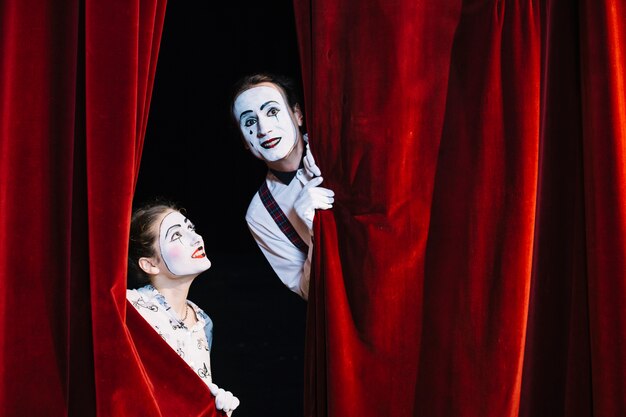 Smiling female mime artist looking at male mime artist peeking from curtain
