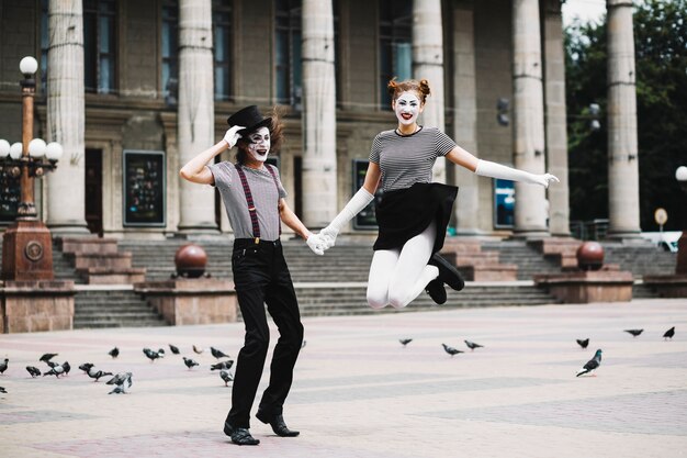 smiling female mime holding male mime's hand jumping in front of building