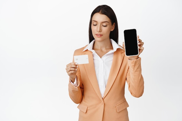 웃는 여성 사무원은 휴대폰 화면과 흰색 배경 위에 서 있는 은행 신청서를 추천하는 신용 카드를 보여줍니다.