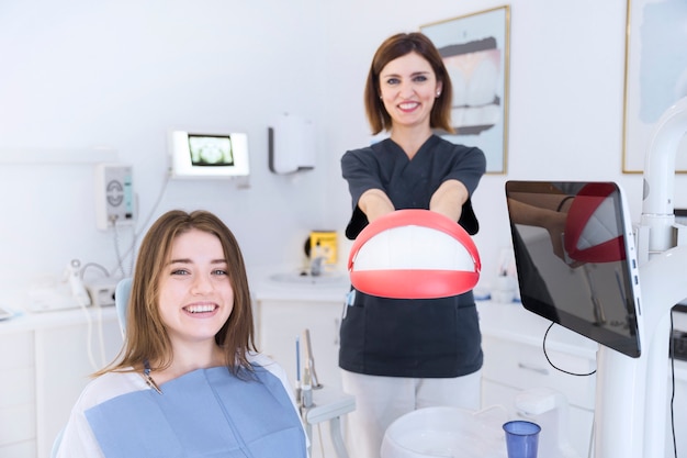 Free photo smiling female patient in front of dentist holding teeth model at clinic