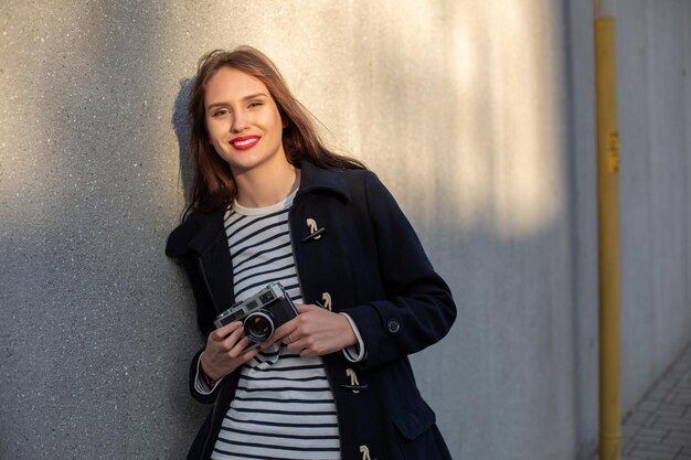 Улыбающаяся женщина-фотограф в куртке стоит перед стеной и готова сделать новое фото. Очаровательная молодая брюнетка в модном наряде позирует на фоне бетонной стены с камерой