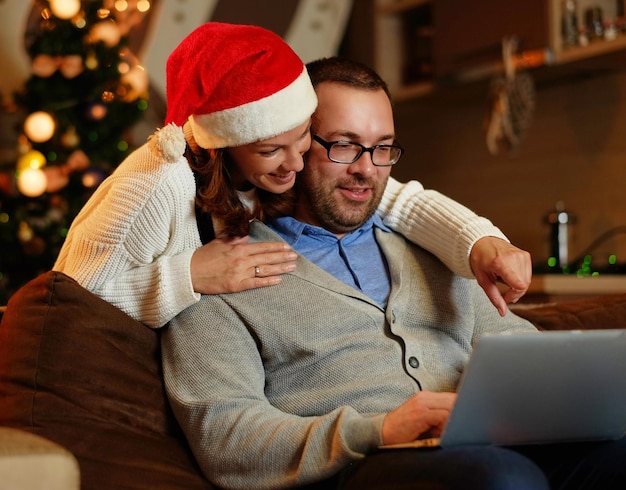 Smiling female in Santa's hat and plump positive male using a laptop on a couch in a room with Christmas decoration.