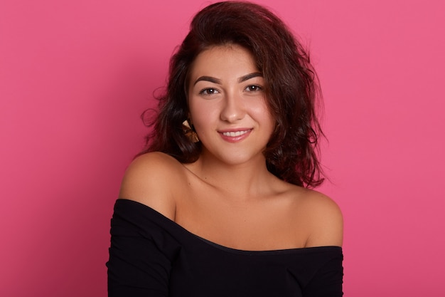 Smiling female with dark wavy hair wearing black outfit,  with glad facial expression, looks confident, posing isolated over pink wall.