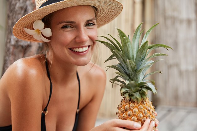 Smiling female with perfect slim body, tanned skin, wears straw hat, holds pineapple