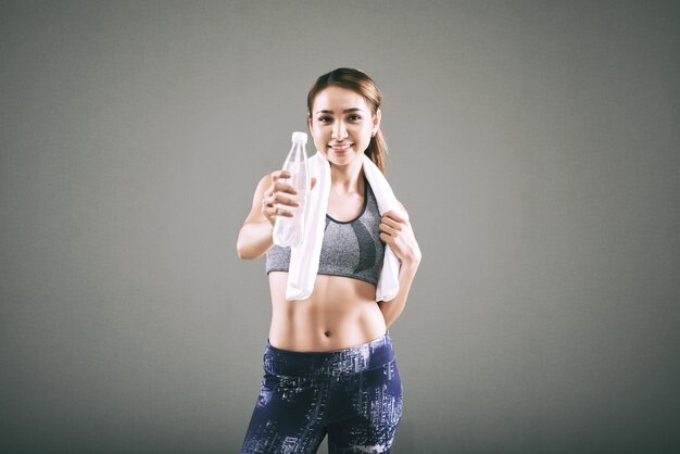 Smiling fit Asian woman in sportswear, with towel on shoulders, holding out bottle of water