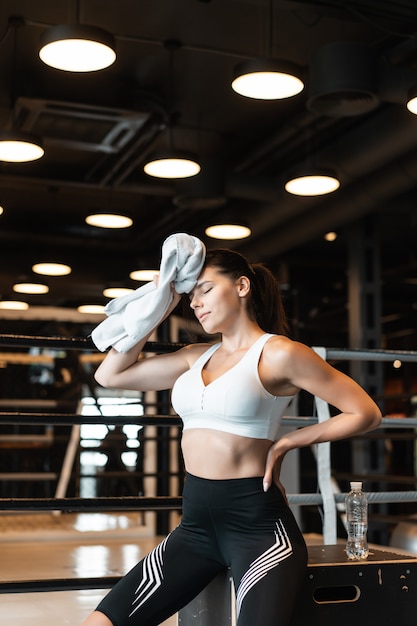 Smiling fit girl holding towel and taking rest in gym. Girl wipes sweat with a towel